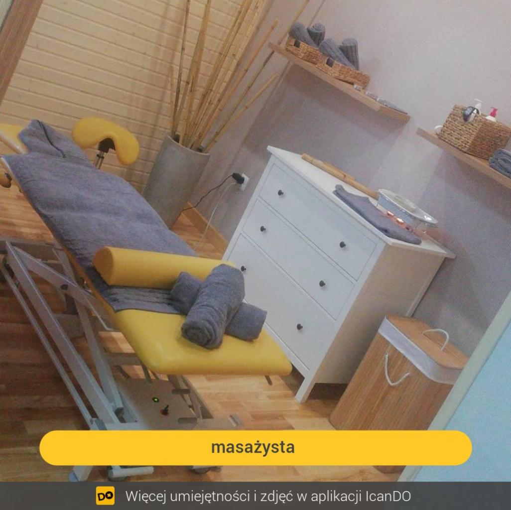 Przygotowane stanowisko do zabiegów: żółty stół z szarą narzutą i zwiniętymi ręcznikami, biała komoda z podgrzewaczem do wosku i bambusowy kosz na pranie w tle.