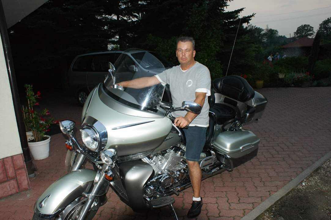 Mężczyzna opiera rękę na srebrnym motocyklu turystycznym na tle domu i zaparkowanego auta. Widoczne detale chromowane i duża szyba motocykla.