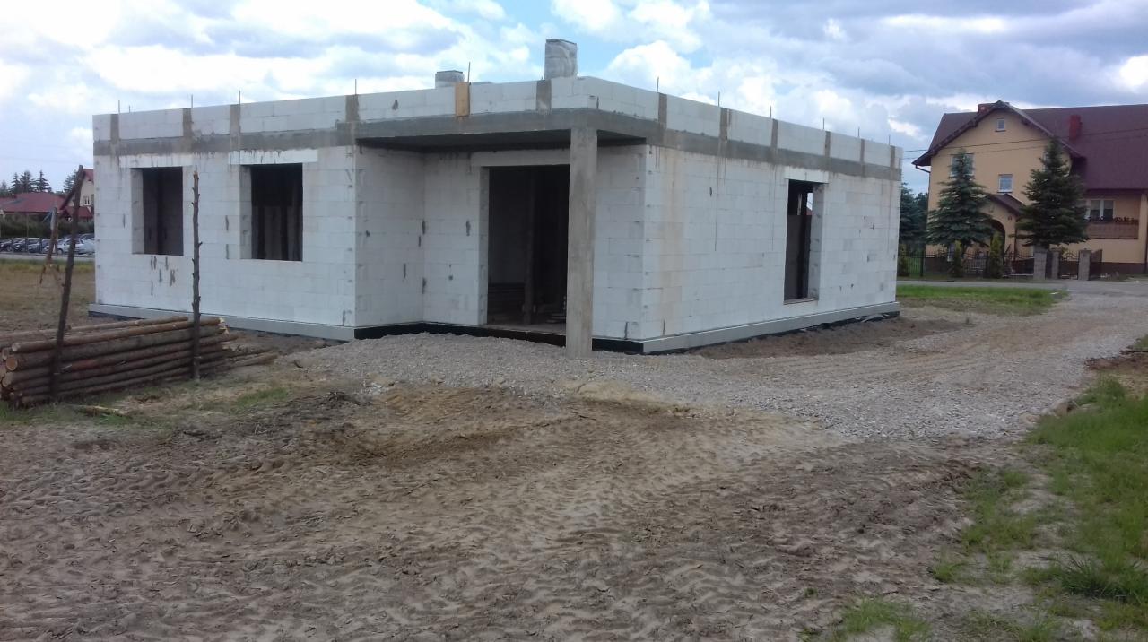 Surowy stan budynku z białych bloczków z otworami okiennymi i wejściowymi, widoczne drewno budowlane obok, w tle dom jednorodzinny i pochmurne niebo.