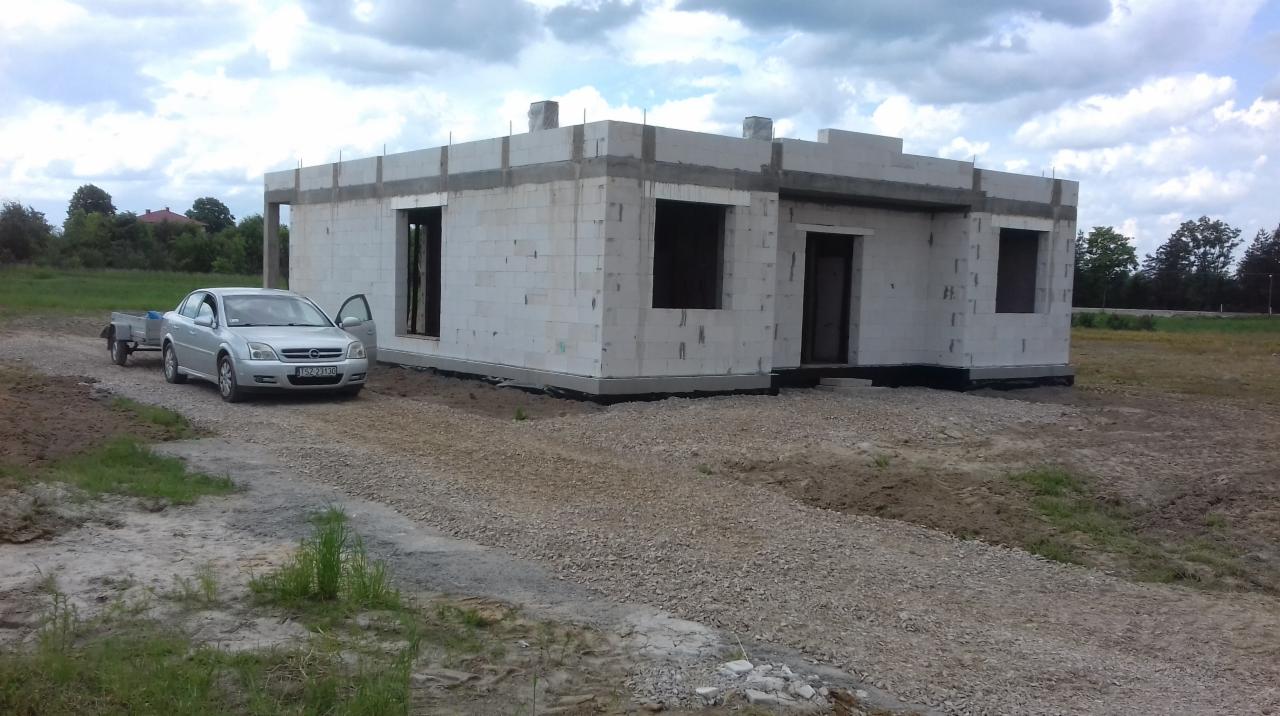 Niedokończony budynek z białych bloczków z otwartymi otworami okiennymi i drzwiami, zaparkowany srebrny samochód osobowy z otwartymi drzwiami i przyczepką na żwirowej drodze.