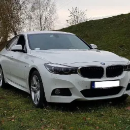 Białe BMW Gran Turismo zaparkowane na trawiastym wzgórzu, widok z przodu, tablica rejestracyjna częściowo zasłonięta.