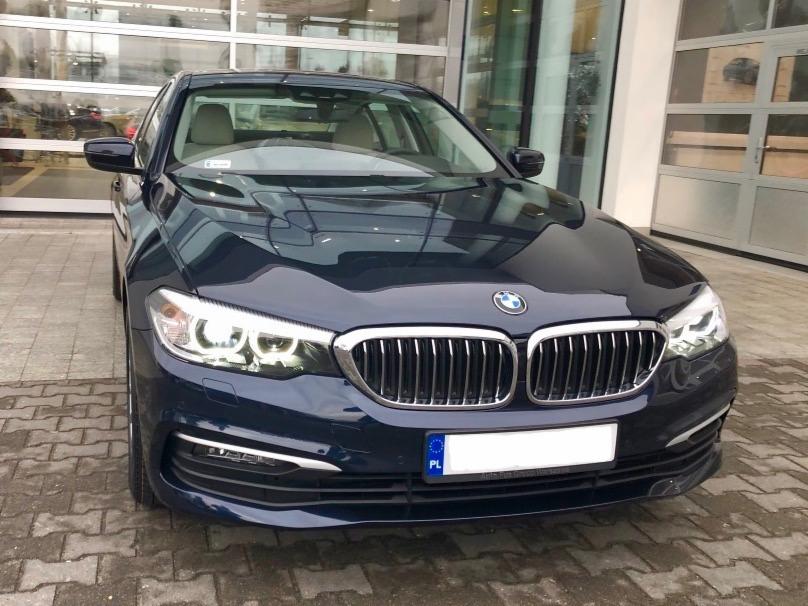Granatowy samochód osobowy marki BMW serii 5, zaparkowany przed przeszklonym salonem samochodowym, z widoczną polską tablicą rejestracyjną.
