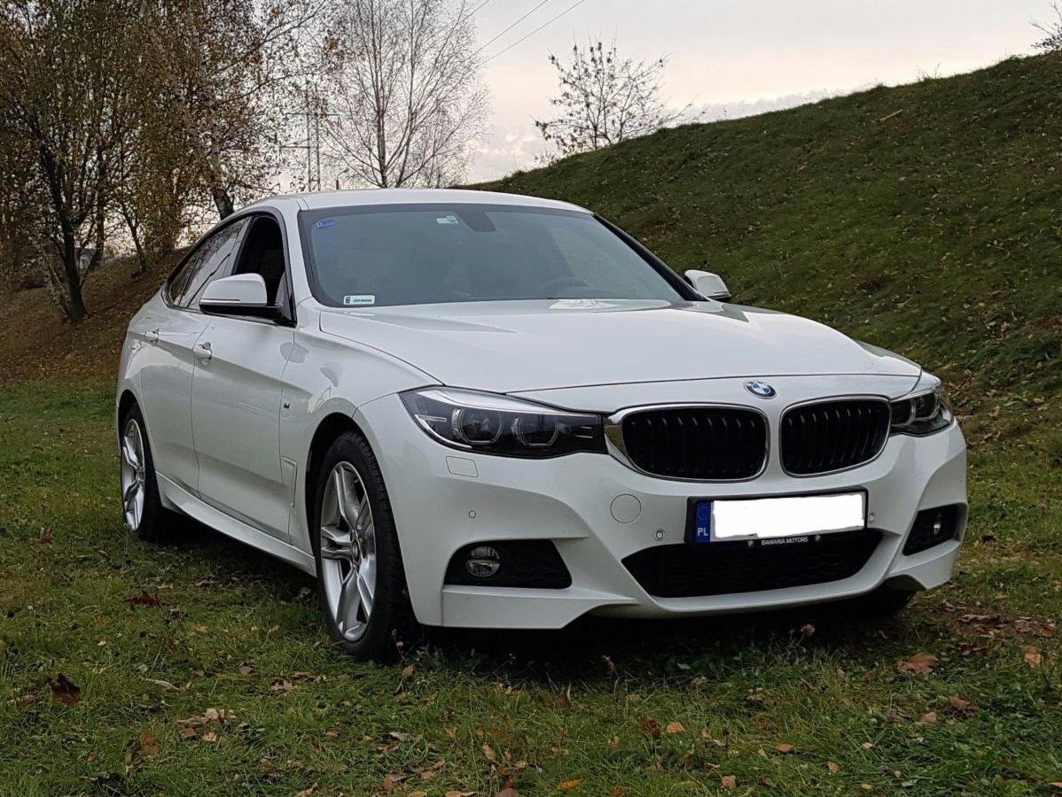 Białe BMW Gran Turismo zaparkowane na trawiastym wzgórzu, widok z przodu, tablica rejestracyjna częściowo zasłonięta.
