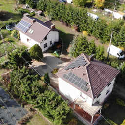 PB-TECH Piotr Borkowski - Widok z lotu ptaka na dwa domy jednorodzinne z brązowymi dachami, na których zamontowane są panele fotowoltaiczne. Biały van zaparkowany w pobliżu jednego z domów.