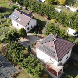 Widok z lotu ptaka na dwa domy jednorodzinne z brązowymi dachami, na których zamontowane są panele fotowoltaiczne. Biały van zaparkowany w pobliżu jednego z domów.