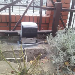 Zamontowany automat do bramy przesuwnej, widoczne okablowanie w ziemi, obok roślina lawendy i fragment ogrodzenia z ceglaną podstawą.