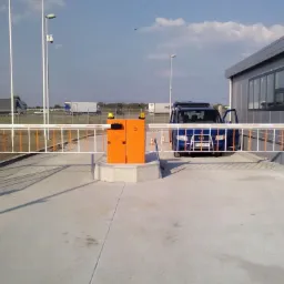 Otwarta szlabanowa brama wjazdowa z pomarańczową obudową automatu, niebieski samochód osobowy z otwartymi drzwiami po prawej stronie, w tle budynek ochrony i ciężarówki.