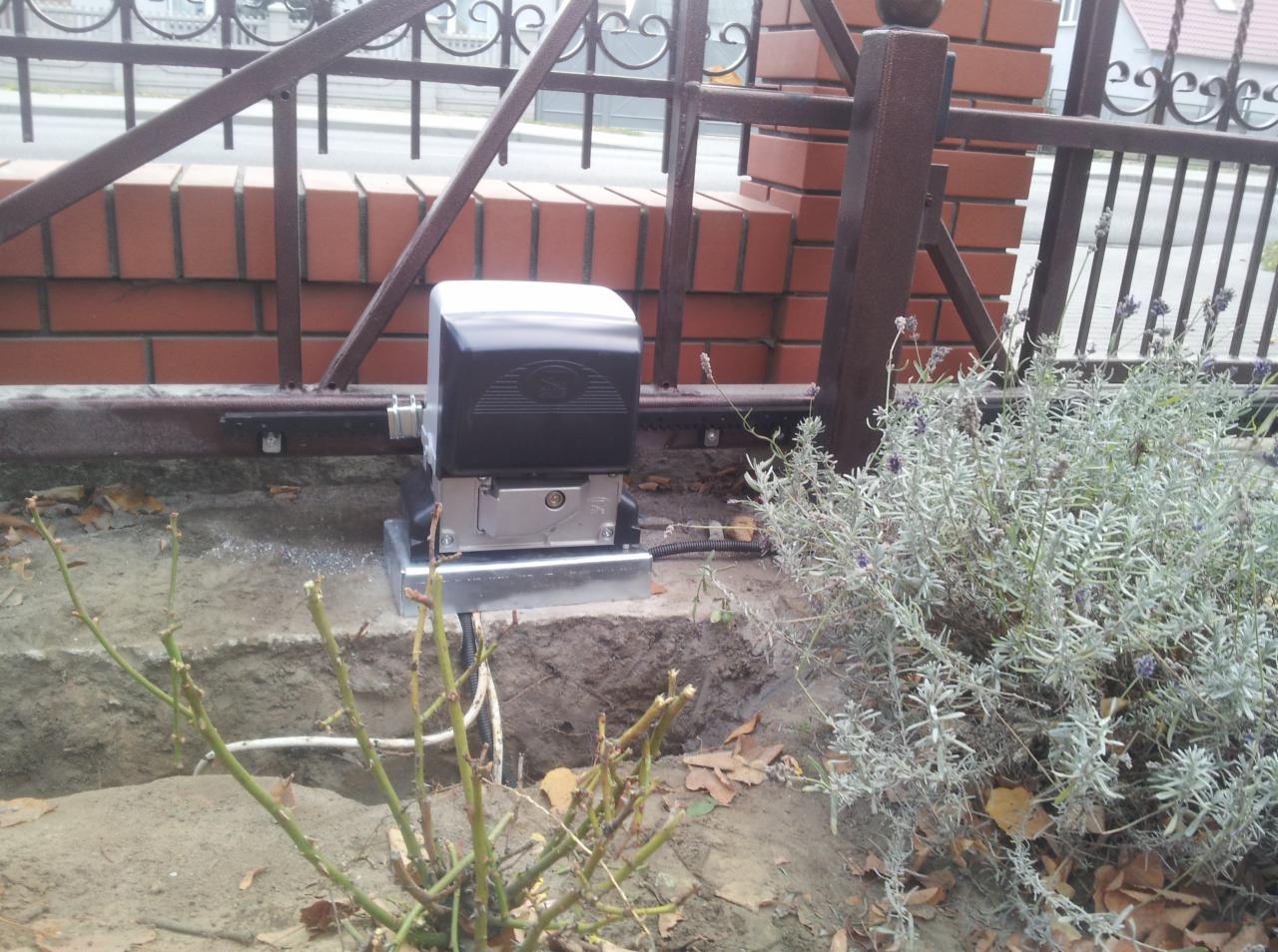 Zamontowany automat do bramy przesuwnej, widoczne okablowanie w ziemi, obok roślina lawendy i fragment ogrodzenia z ceglaną podstawą.