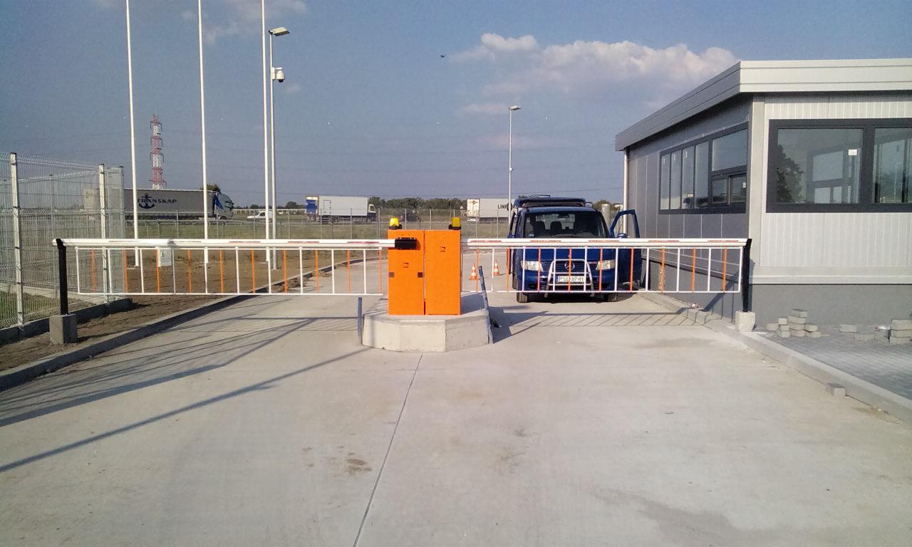 Otwarta szlabanowa brama wjazdowa z pomarańczową obudową automatu, niebieski samochód osobowy z otwartymi drzwiami po prawej stronie, w tle budynek ochrony i ciężarówki.