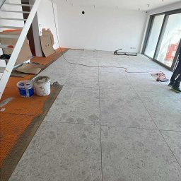 Glory - Układanie szarych, kwadratowych płytek podłogowych w jasnym wnętrzu. Widoczne narzędzia glazurnicze i fragment schodów. Częściowo ułożona podłoga na pomarańczowej macie.