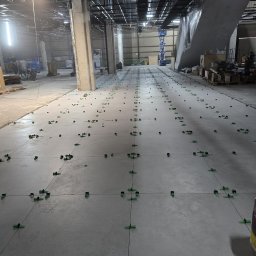 Glory - Układanie płytek w dużej hali. Widoczne krzyżyki dystansowe i sznurki wyznaczające linie. W tle filary i instalacje pod sufitem. Etap prac wykończeniowych.