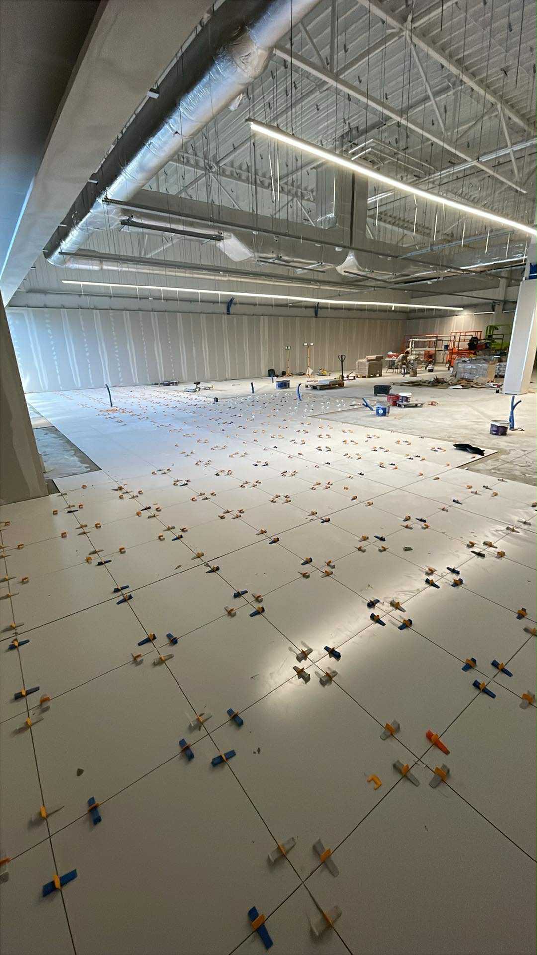 Układanie jasnych płytek w dużym pomieszczeniu z widocznymi krzyżykami dystansowymi. W tle widoczne instalacje pod sufitem i sprzęt budowlany.