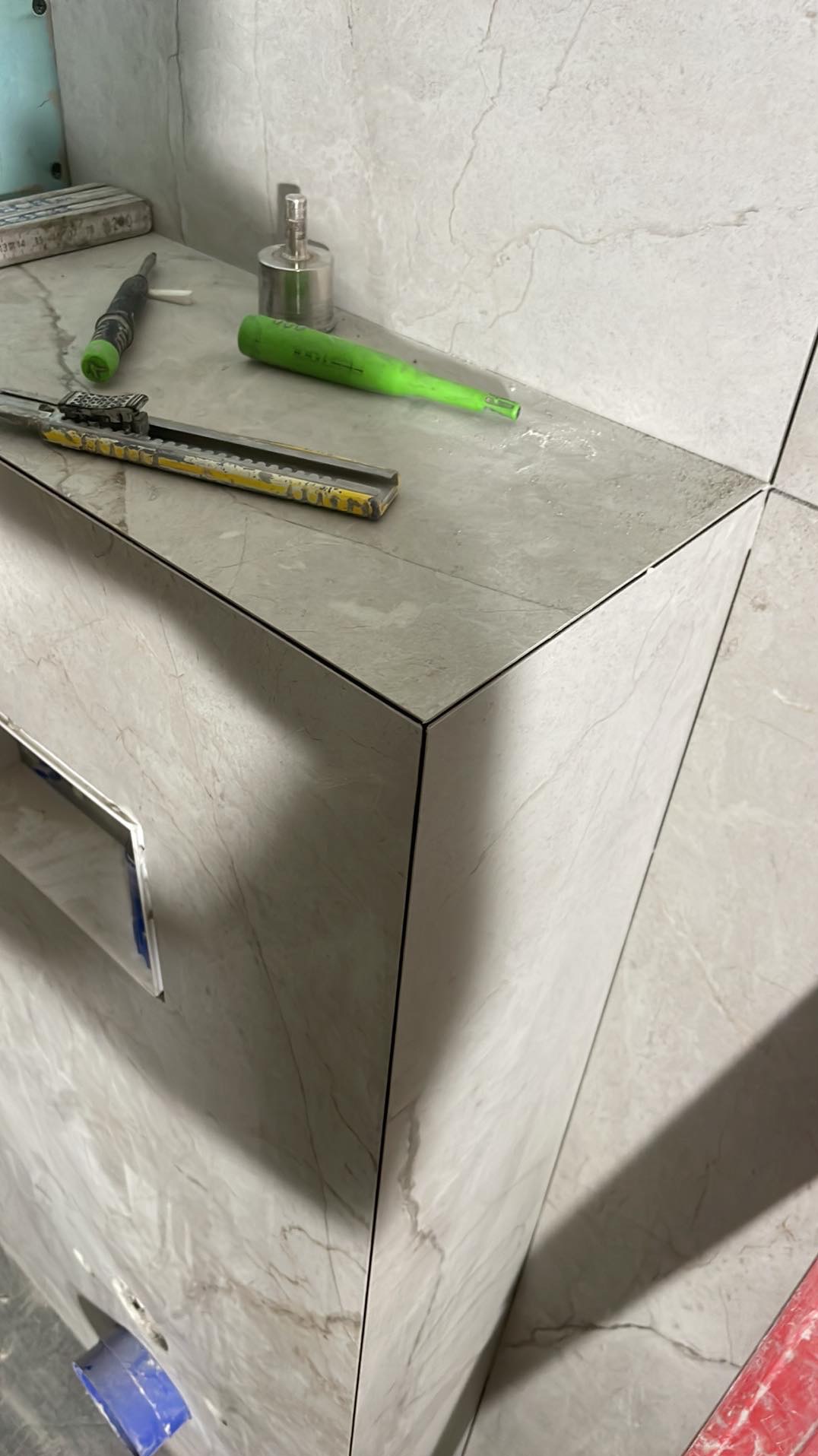 Wykończona zabudowa z płytek imitujących marmur, z widocznymi narzędziami glazurniczymi na blacie. Precyzyjne łączenia płytek, minimalistyczny design.