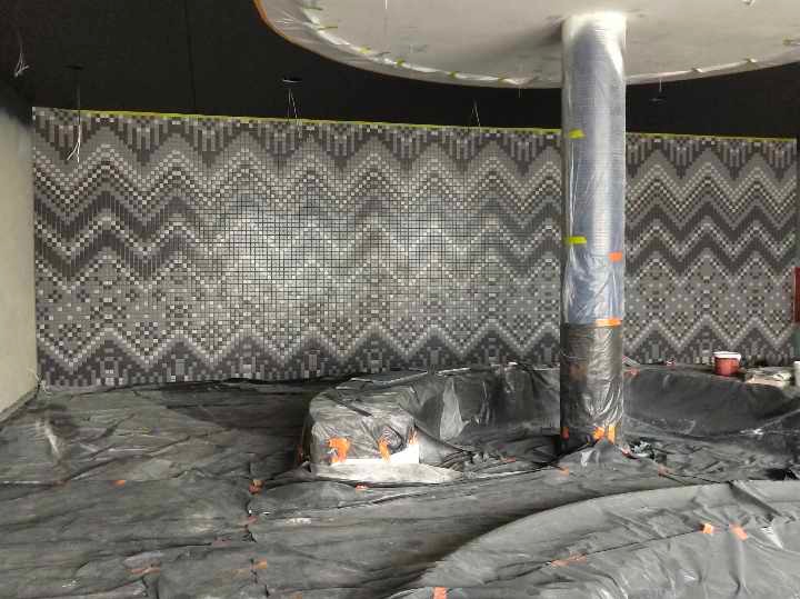 Mozaika w zygzaki na ścianie w trakcie montażu. Podłoga zabezpieczona folią. Kolumna owinięta folią ochronną. Widoczne narzędzia i materiały budowlane.