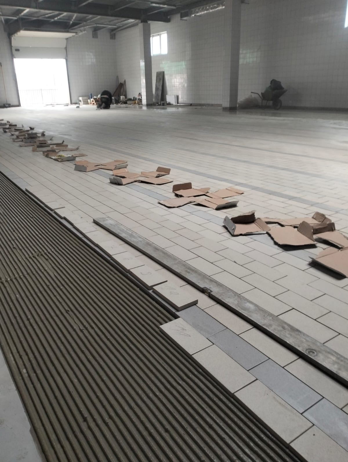 Układanie płytek podłogowych w dużym pomieszczeniu. Widoczna warstwa kleju, płytki i kartony zabezpieczające. Perspektywa z bliska, ukazująca detale pracy.