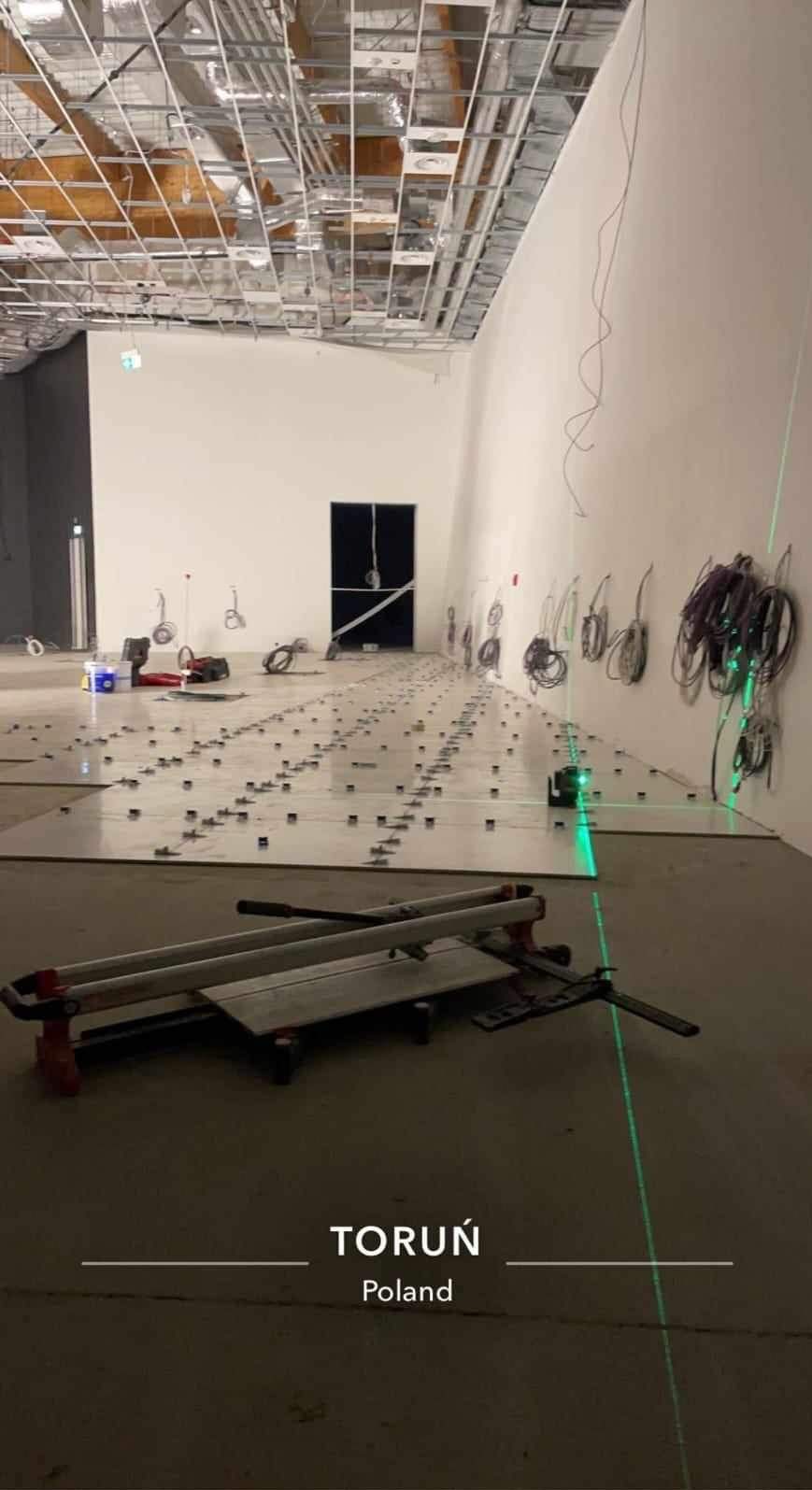 Układanie płytek podłogowych w dużym pomieszczeniu z użyciem poziomicy laserowej i narzędzi glazurniczych. Widoczne płytki, klej i system poziomowania.