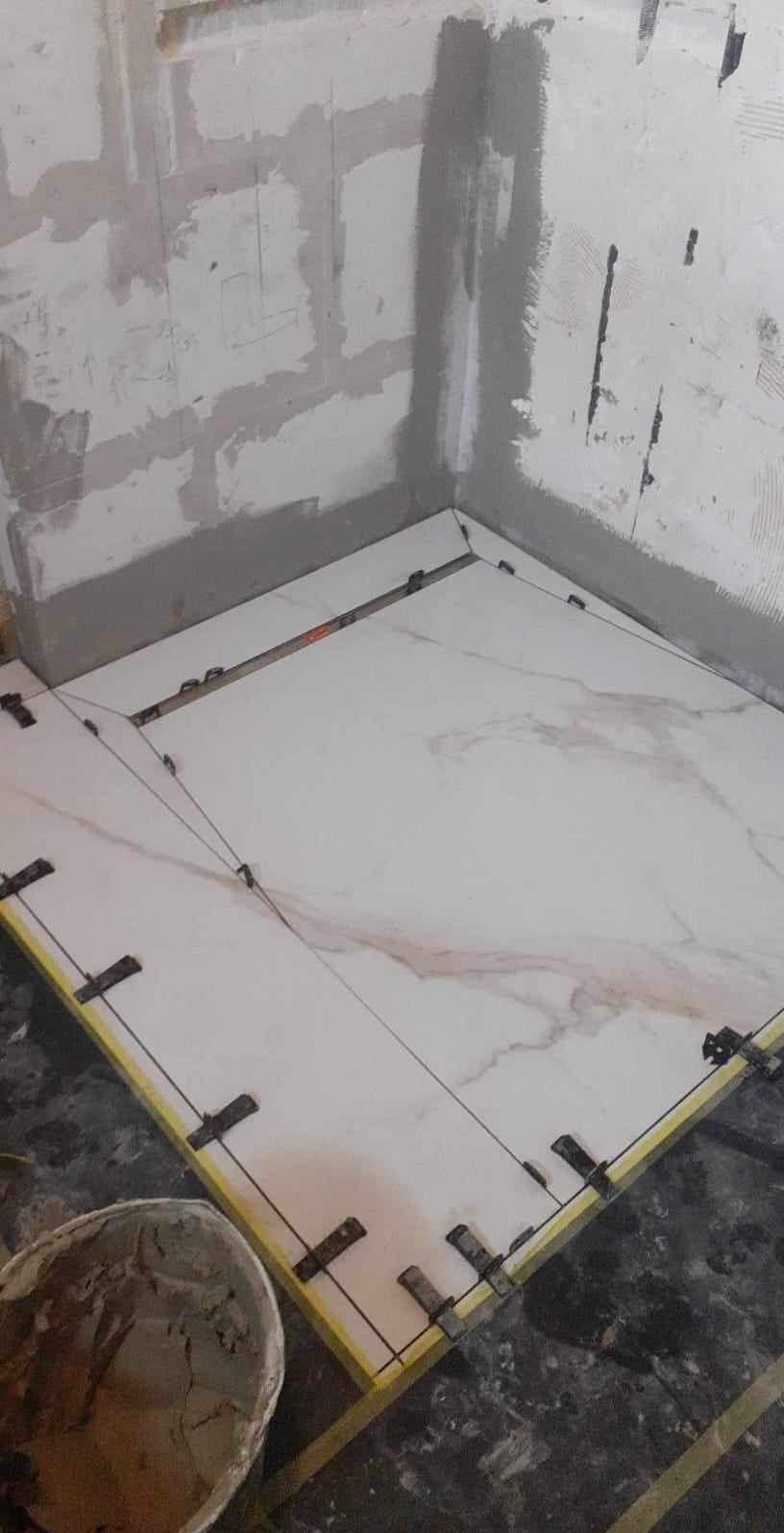 Układanie płytek imitujących marmur w rogu pomieszczenia. Widoczne klipsy poziomujące i wiadro z zaprawą. Ściany surowe, przygotowane do dalszych prac wykończeniowych.