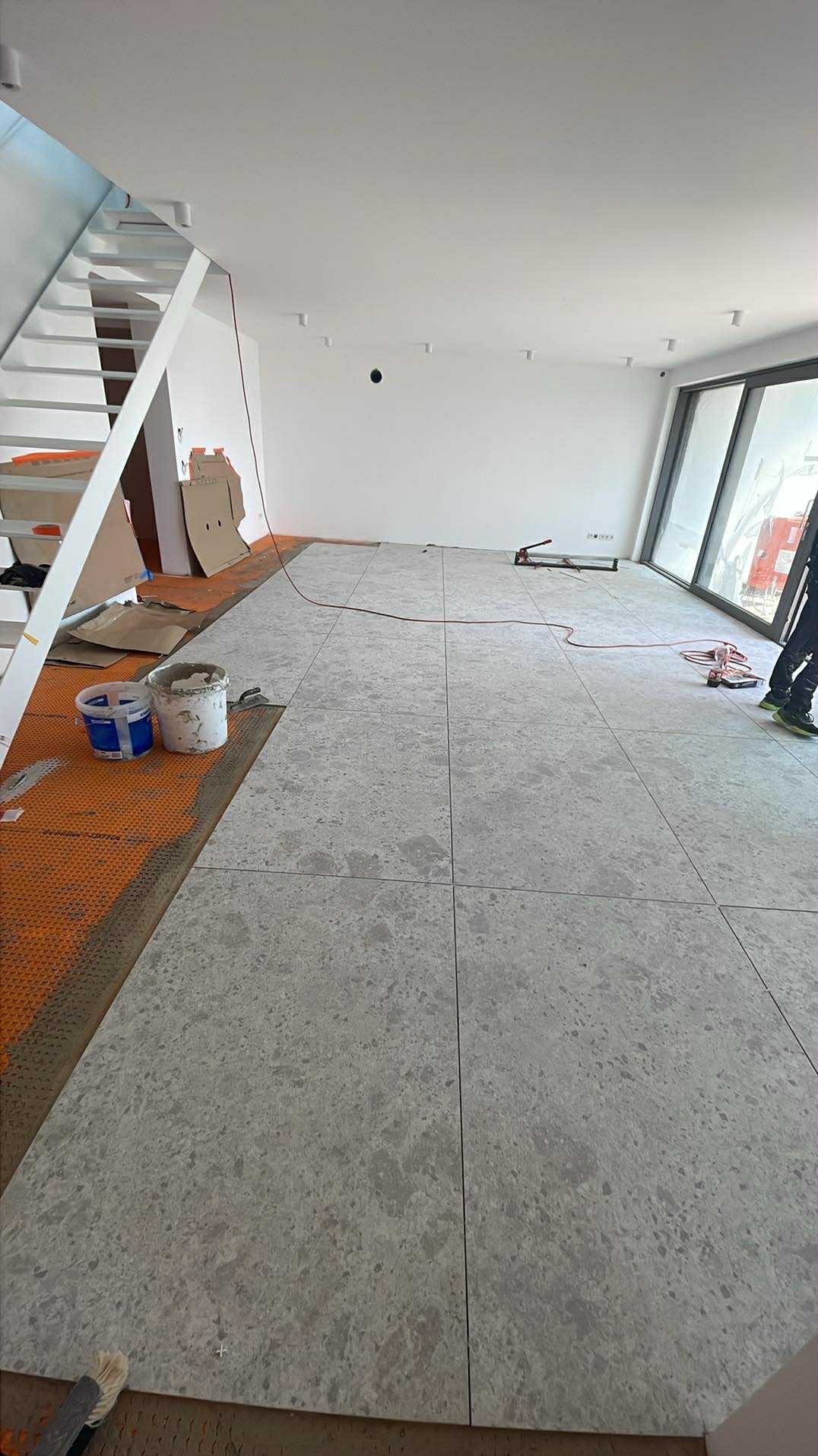 Układanie szarych, kwadratowych płytek podłogowych w jasnym wnętrzu. Widoczne narzędzia glazurnicze i fragment schodów. Częściowo ułożona podłoga na pomarańczowej macie.