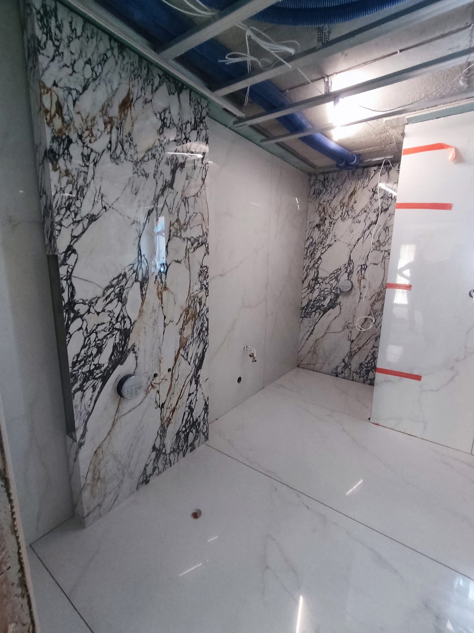 Wnętrze łazienki w trakcie wykańczania: marmurowe płytki na ścianach, biała podłoga, widoczne instalacje pod sufitem. Częściowo wykończona kabina prysznicowa.