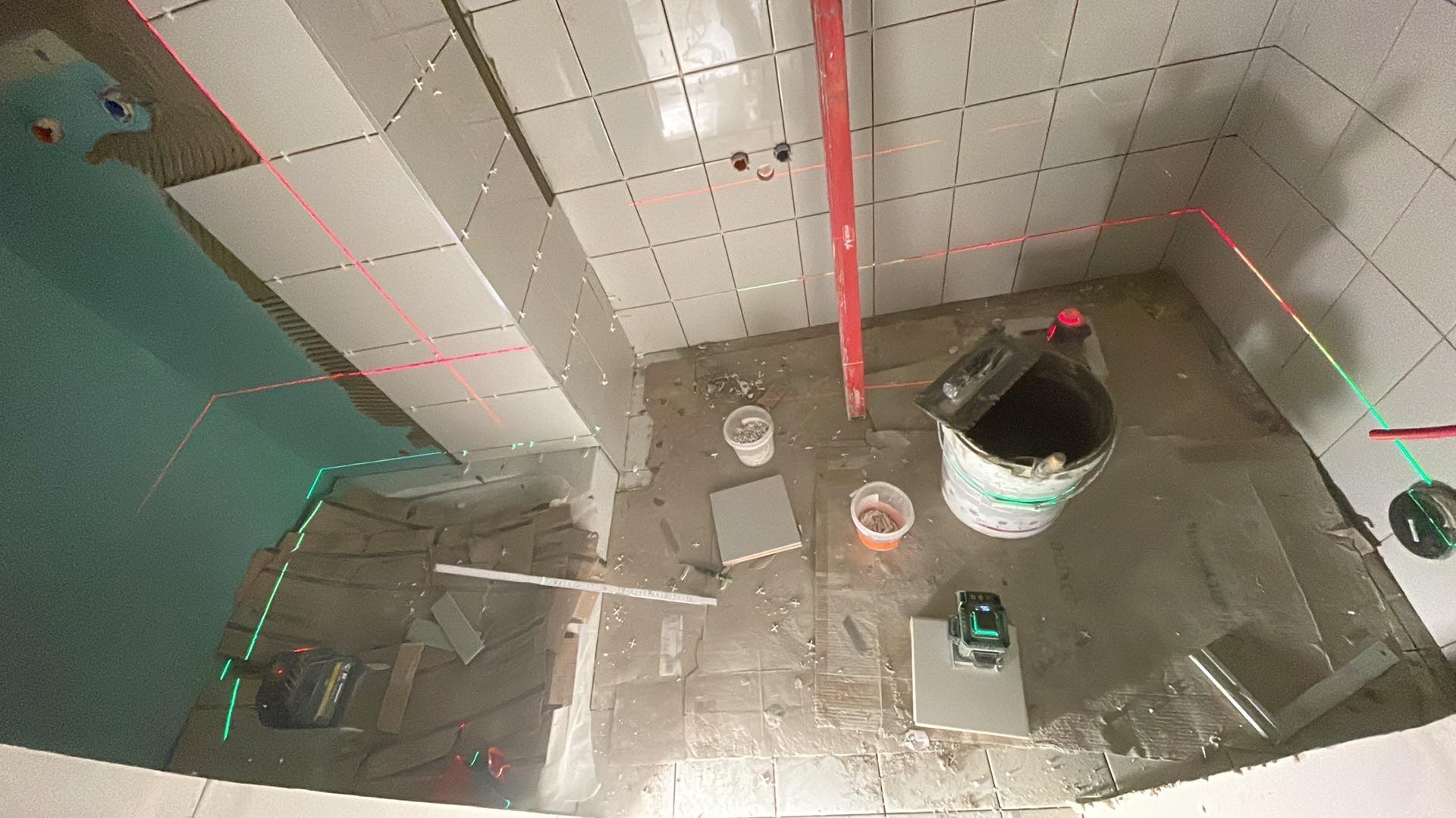 Układanie płytek w łazience z użyciem lasera krzyżowego. Widoczne narzędzia, wiadro z klejem i częściowo ułożone płytki na ścianach. Prace w toku, surowy stan.