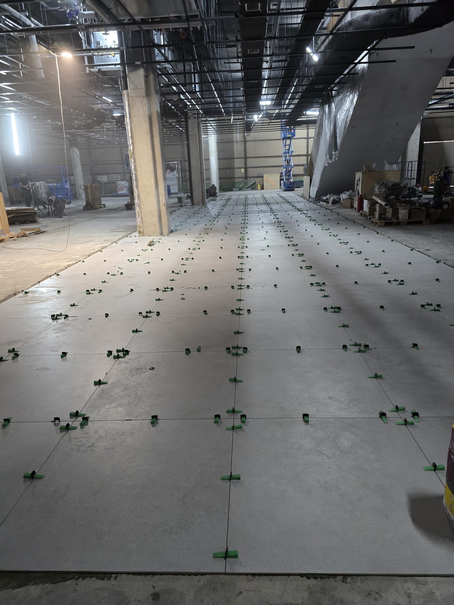 Układanie płytek w dużej hali. Widoczne krzyżyki dystansowe i sznurki wyznaczające linie. W tle filary i instalacje pod sufitem. Etap prac wykończeniowych.