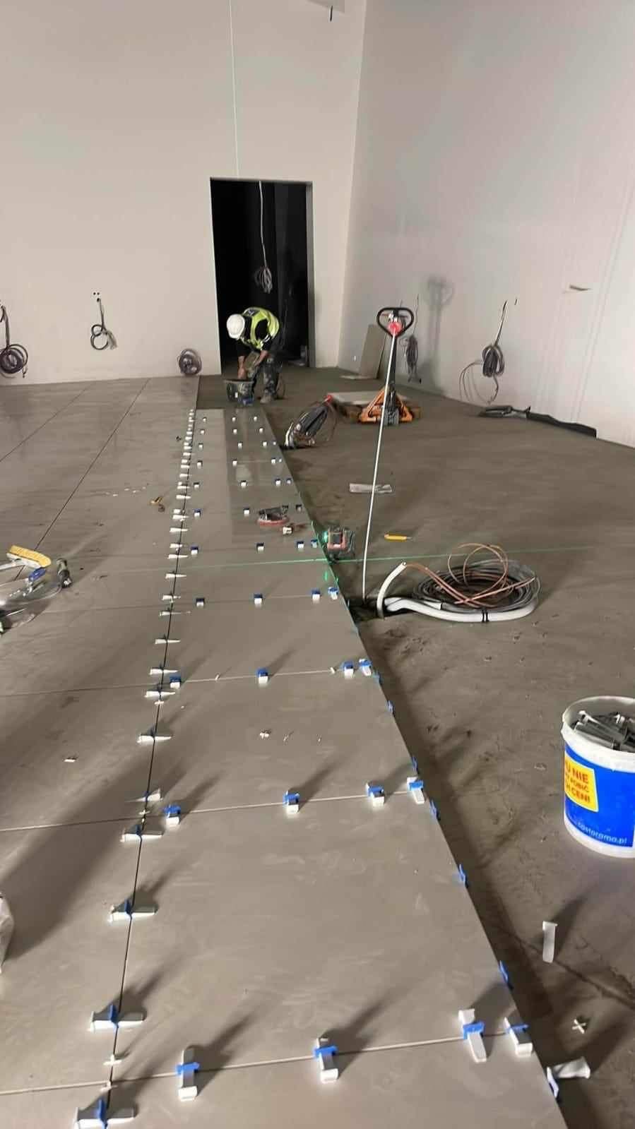 Układanie płytek podłogowych w dużym pomieszczeniu, widoczne narzędzia glazurnicze i pracownik w tle. Użyto poziomicy laserowej do precyzyjnego montażu.