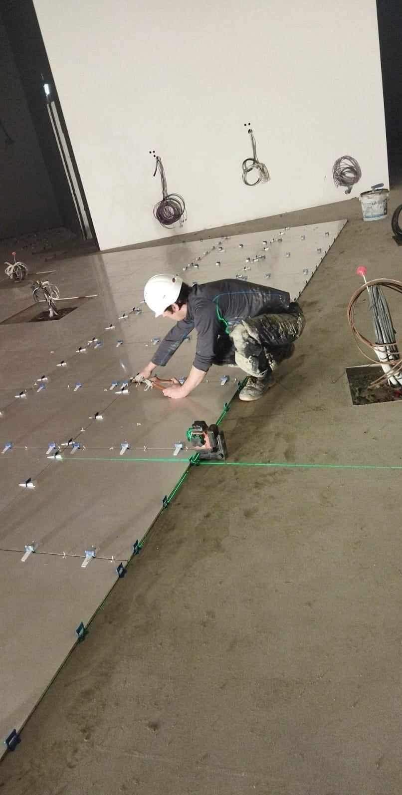 Układanie płytek podłogowych z użyciem poziomicy laserowej i systemu poziomowania. Widoczne kable i instalacje w tle. Fachowiec w kasku podczas pracy.