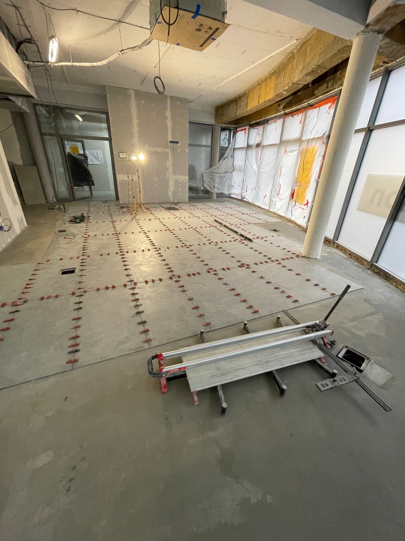 Przygotowanie podłoża pod płytki: poziomowanie z użyciem klipsów i krzyżyków. Na pierwszym planie gilotyna do cięcia płytek. Widoczne okna i ściany w trakcie prac.