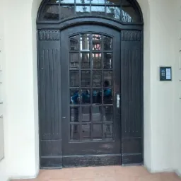 Ciemne, drewniane drzwi wejściowe z łukowatym nadprożem i szprosami, widoczne odbicie otoczenia w szybach, obok domofon