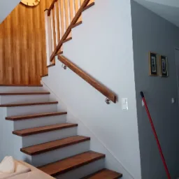 Drewniane schody z białymi podstopnicami i drewnianą balustradą z białymi szczeblami, widok z dołu, fragment sofy w dolnym lewym rogu, na ścianie dwa obrazy w złotych ramach.