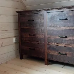 Ciemnobrązowa komoda z sześcioma szufladami i czarnymi uchwytami, ustawiona w pomieszczeniu wykończonym jasnym drewnem, widoczny fragment białej tkaniny po prawej stronie.