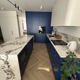 Marcin Pepliński - Nowoczesna kuchnia z wyspą i zabudową w odcieniach bieli i granatu, blaty z kamienia z wyraźnym wzorem, parkiet w jodełkę. Widok na salon w tle.