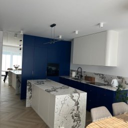 Marcin Pepliński - Nowoczesna kuchnia z wyspą i blatem z marmuru, szafki w odcieniach bieli i granatu, parkiet w jodełkę. Minimalistyczne oświetlenie i okrągły stół dodają elegancji.