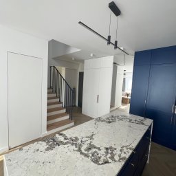 Marcin Pepliński - Wyspa kuchenna z blatem z kamienia we wzory, zabudowa szaf w bieli i granacie, schody z drewnianymi stopniami we Wrocławiu. Nowoczesne oświetlenie.