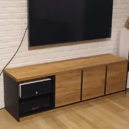 Nowoczesna szafka RTV w odcieniach dębu i czerni z widocznym sprzętem audio, zawieszona pod dużym telewizorem na ścianie z białej cegły.