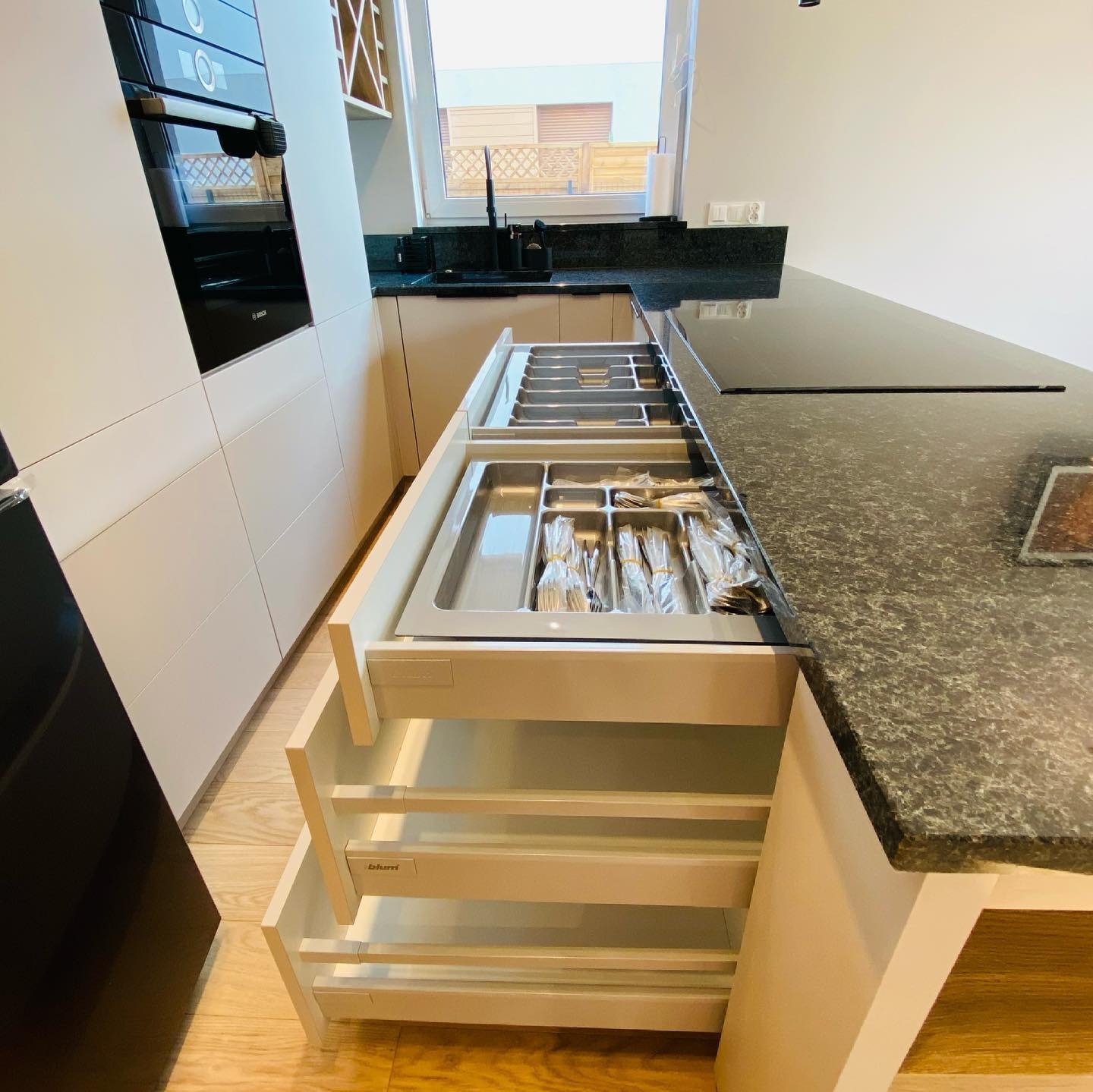 Otwarta szuflada z wkładem na sztućce w nowoczesnej kuchni z czarnym blatem i białymi frontami, widoczny system szuflad Blum.
