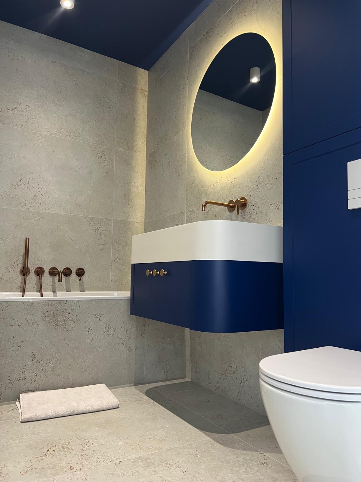 Nowoczesna łazienka z umywalką w kolorze granatowo-białym, okrągłym lustrem z podświetleniem LED i baterią w kolorze miedzianym. Granatowy sufit i zabudowa.
