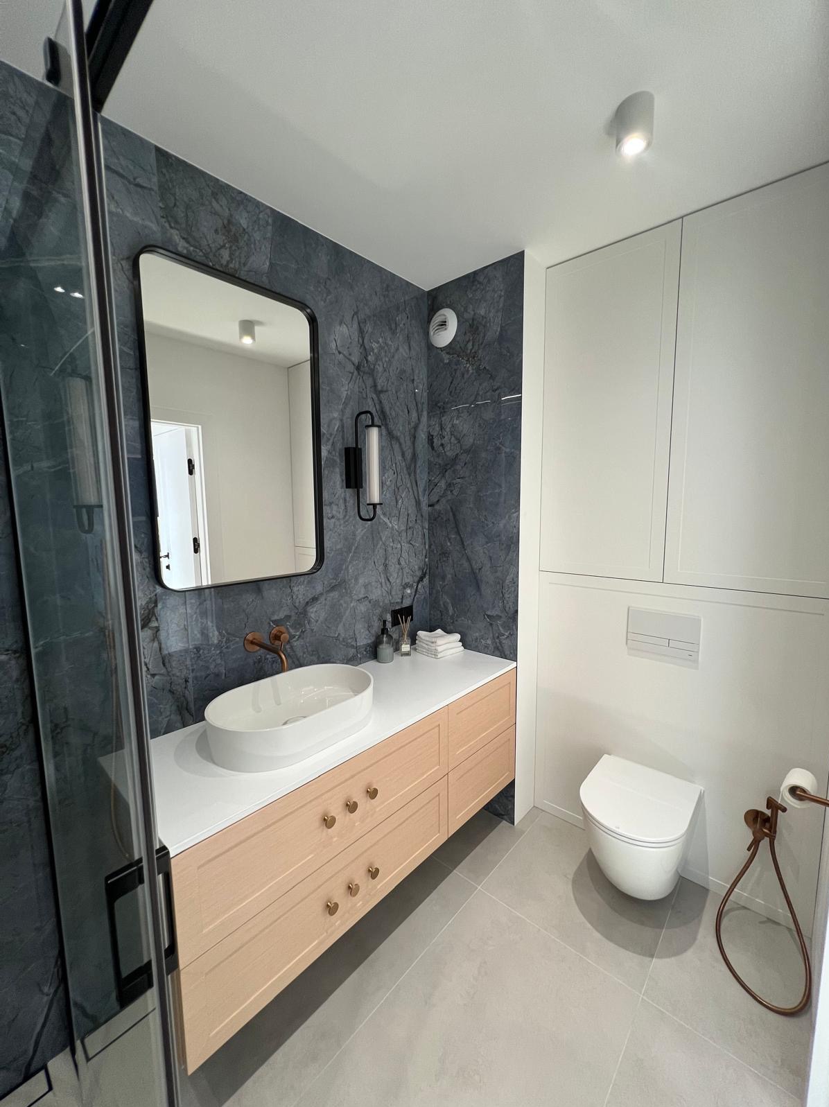 Nowoczesna łazienka z umywalką nablatową, szafką z jasnego drewna i marmurową ścianą. Minimalistyczny design z akcentami w kolorze miedzi.