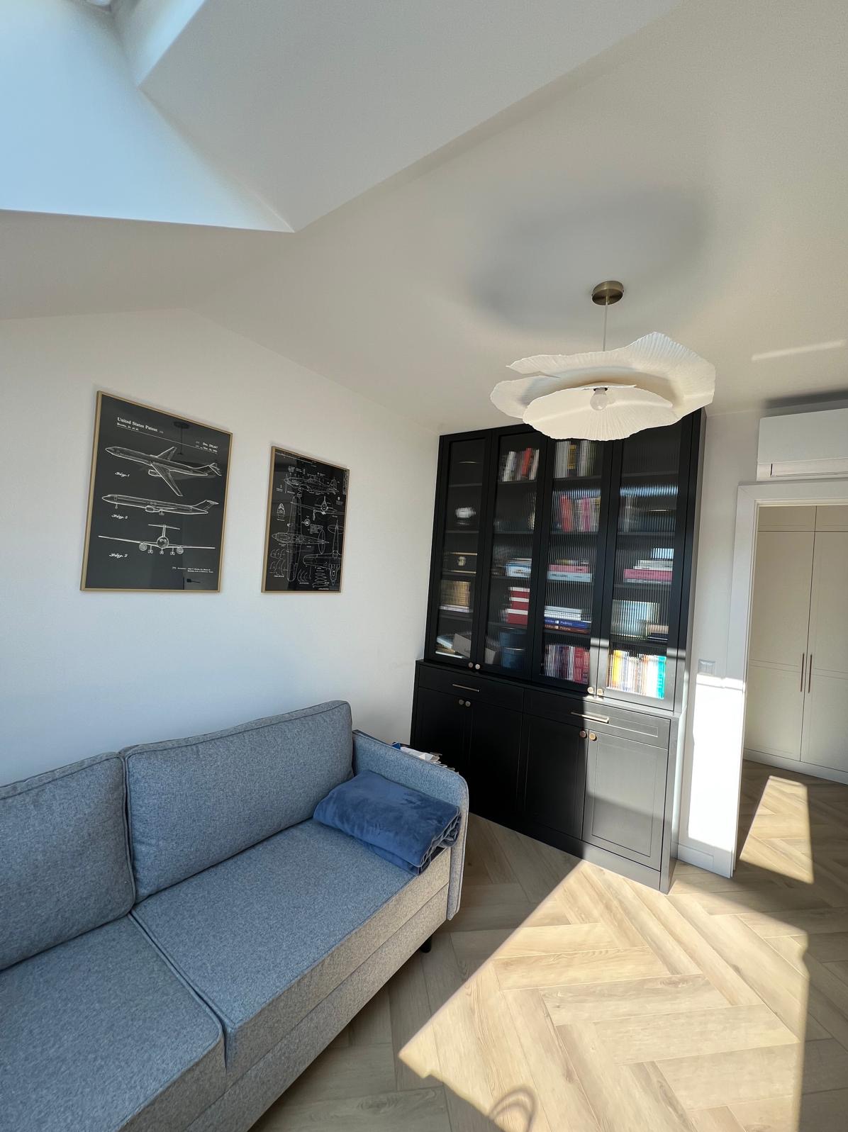 Czarna, przeszklona biblioteczka z książkami w jasnym salonie z szarą sofą i parkietem w jodełkę, oświetlona lampą w kształcie kwiatu. Na ścianie grafiki w ramkach.