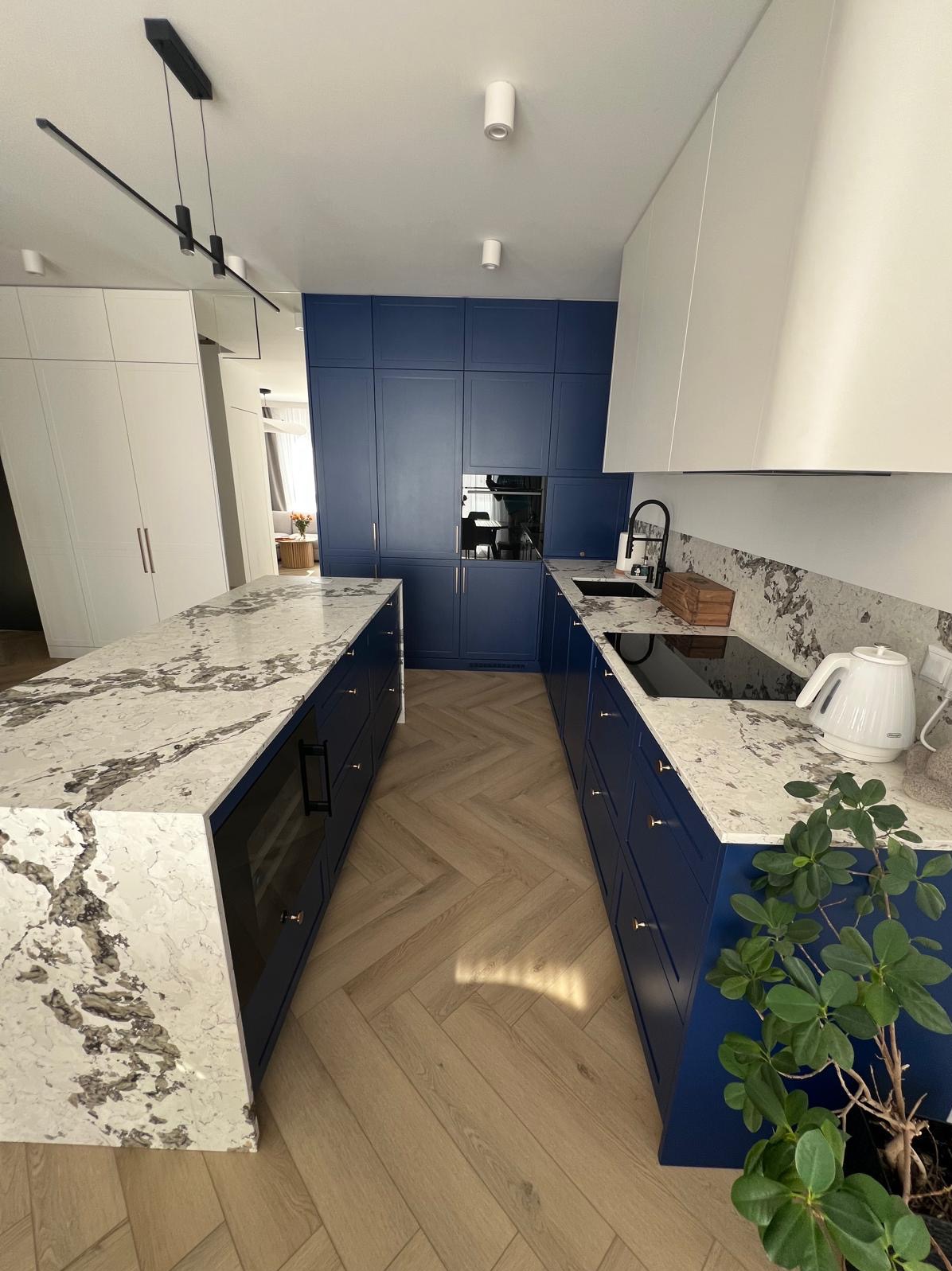 Nowoczesna kuchnia z wyspą i zabudową w odcieniach bieli i granatu, blaty z kamienia z wyraźnym wzorem, parkiet w jodełkę. Widok na salon w tle.