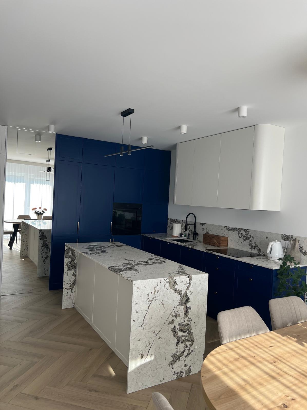 Nowoczesna kuchnia z wyspą i blatem z marmuru, szafki w odcieniach bieli i granatu, parkiet w jodełkę. Minimalistyczne oświetlenie i okrągły stół dodają elegancji.