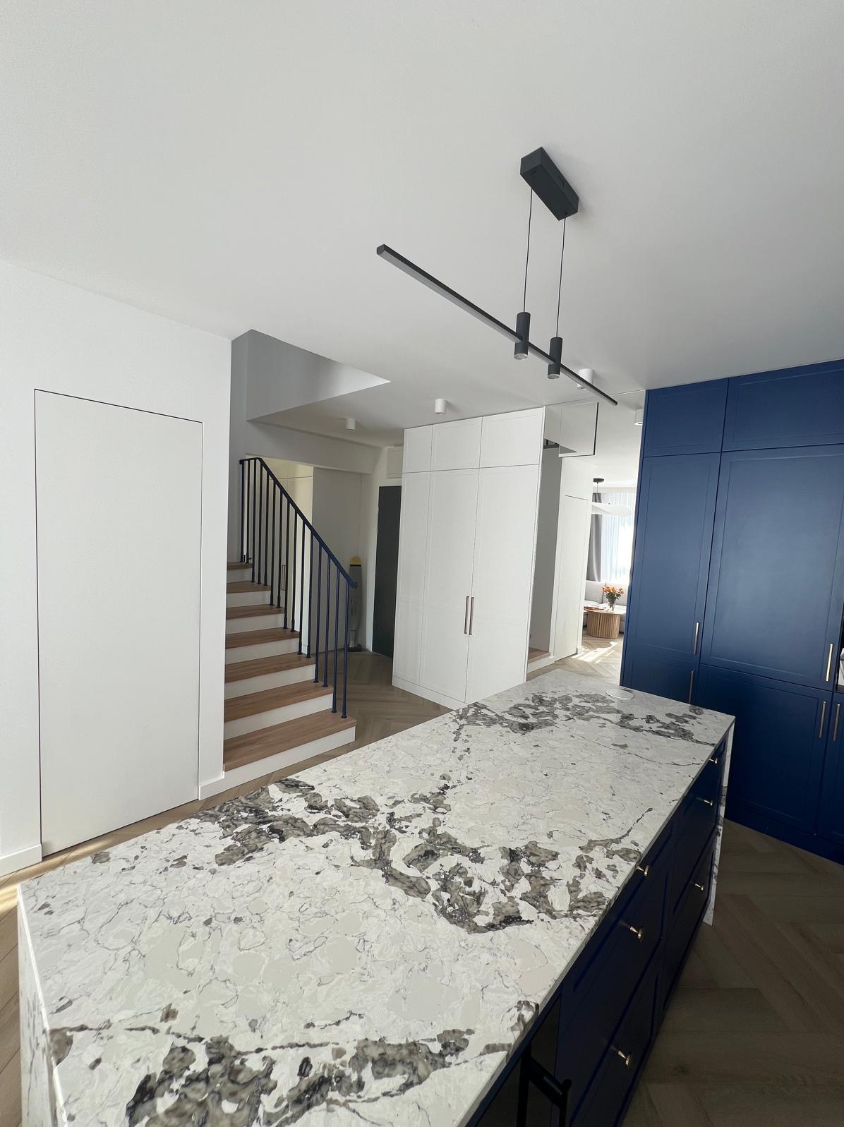 Wyspa kuchenna z blatem z kamienia we wzory, zabudowa szaf w bieli i granacie, schody z drewnianymi stopniami we Wrocławiu. Nowoczesne oświetlenie.