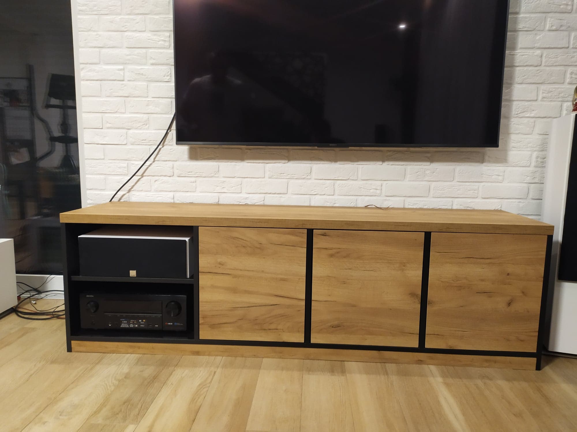 Drewniana szafka RTV z czarnymi elementami, na niej telewizor, na półkach sprzęt audio, ściana z białej cegły.