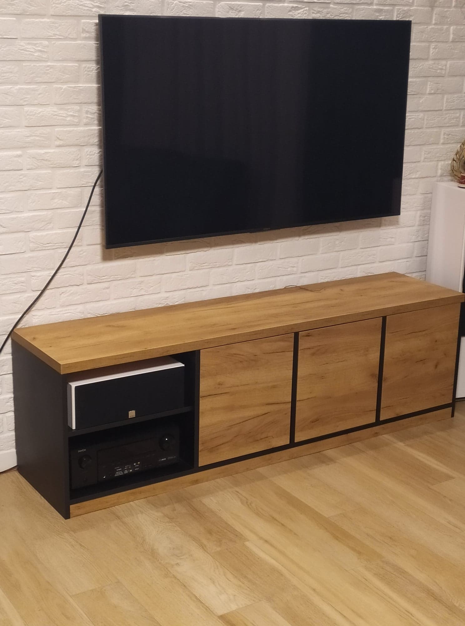 Nowoczesna szafka RTV w odcieniach dębu i czerni z widocznym sprzętem audio, zawieszona pod dużym telewizorem na ścianie z białej cegły.