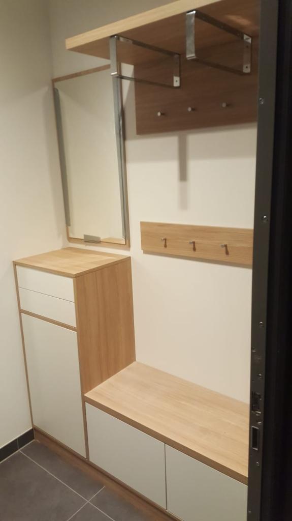 Nowoczesny przedpokój z zabudową: szafka z szufladami, siedzisko z szafką, lustro w cienkiej ramie oraz panel z trzema wieszakami.