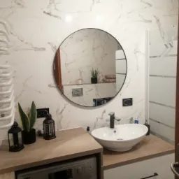 Nowoczesna łazienka z okrągłym lustrem, białą umywalką nablatową, grzejnikiem drabinkowym i marmurowymi płytkami ściennymi, z pralką ukrytą pod blatem.