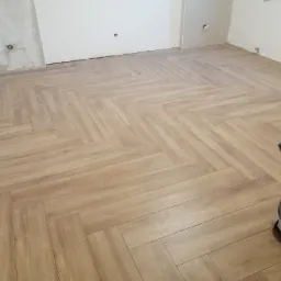 Ułożona podłoga z paneli w jodełkę w jasnym odcieniu drewna, widoczny odkurzacz przemysłowy i surowe ściany przygotowane do malowania.