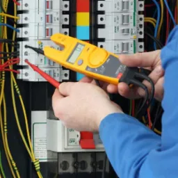 Elektryk w niebieskim kombinezonie mierzy napięcie w skrzynce elektrycznej za pomocą żółtego miernika cęgowego z czarnymi przewodami pomiarowymi i czerwoną sondą, widoczne bezpieczniki i okablowanie.