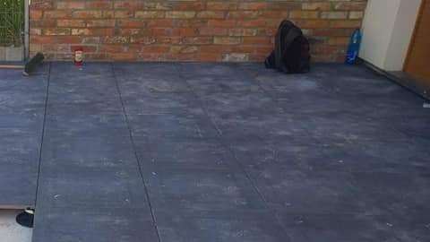 Układanie ciemnej kostki brukowej na tarasie, widoczna część podłoża, ceglana ściana w tle, narzędzia i plecak obok.