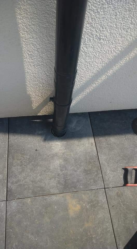 Ciemna rura spustowa na tle szarej ściany i ułożonej nawierzchni z płytek betonowych.